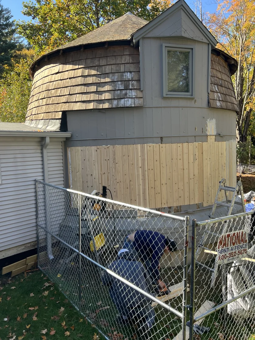 The Silo during siding repairs, with new wooden boards along its curved exterior, a ladder and a worker behind temporary fencing.